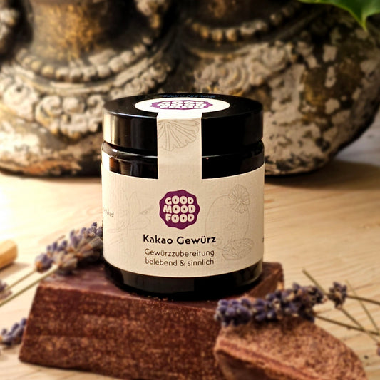 Schraubglas gefüllt mit Kakaogewürz platziert auf Rohkakaoblock, der auf einem Holztisch liegt mit einer Deko-Betonfigur im Hintergrund.